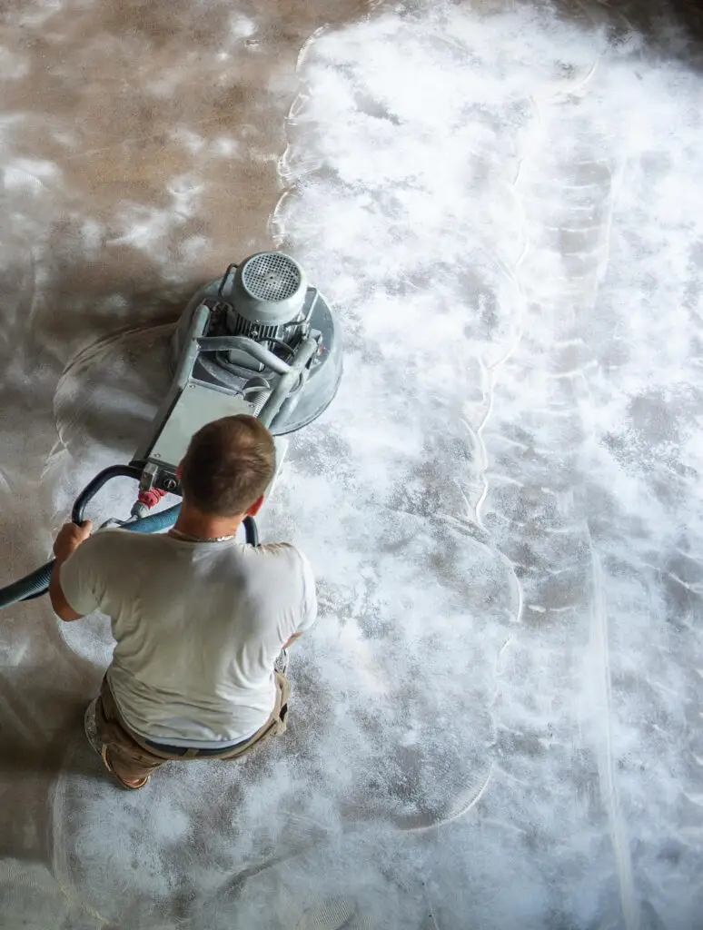 A person operates a floor grinding machine, smoothing a concrete surface, leaving a visible contrast of polished and unpolished areas.