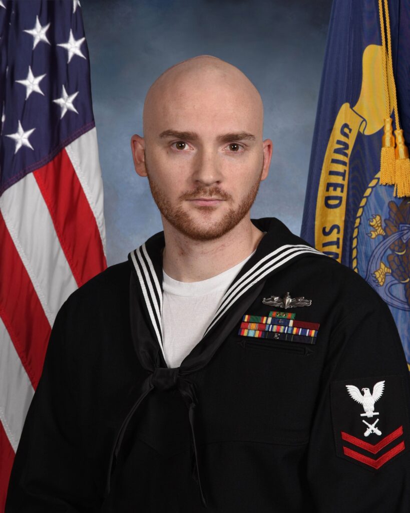 A person in a Navy uniform stands in front of American and state flags, displaying several medals and insignia on their attire.